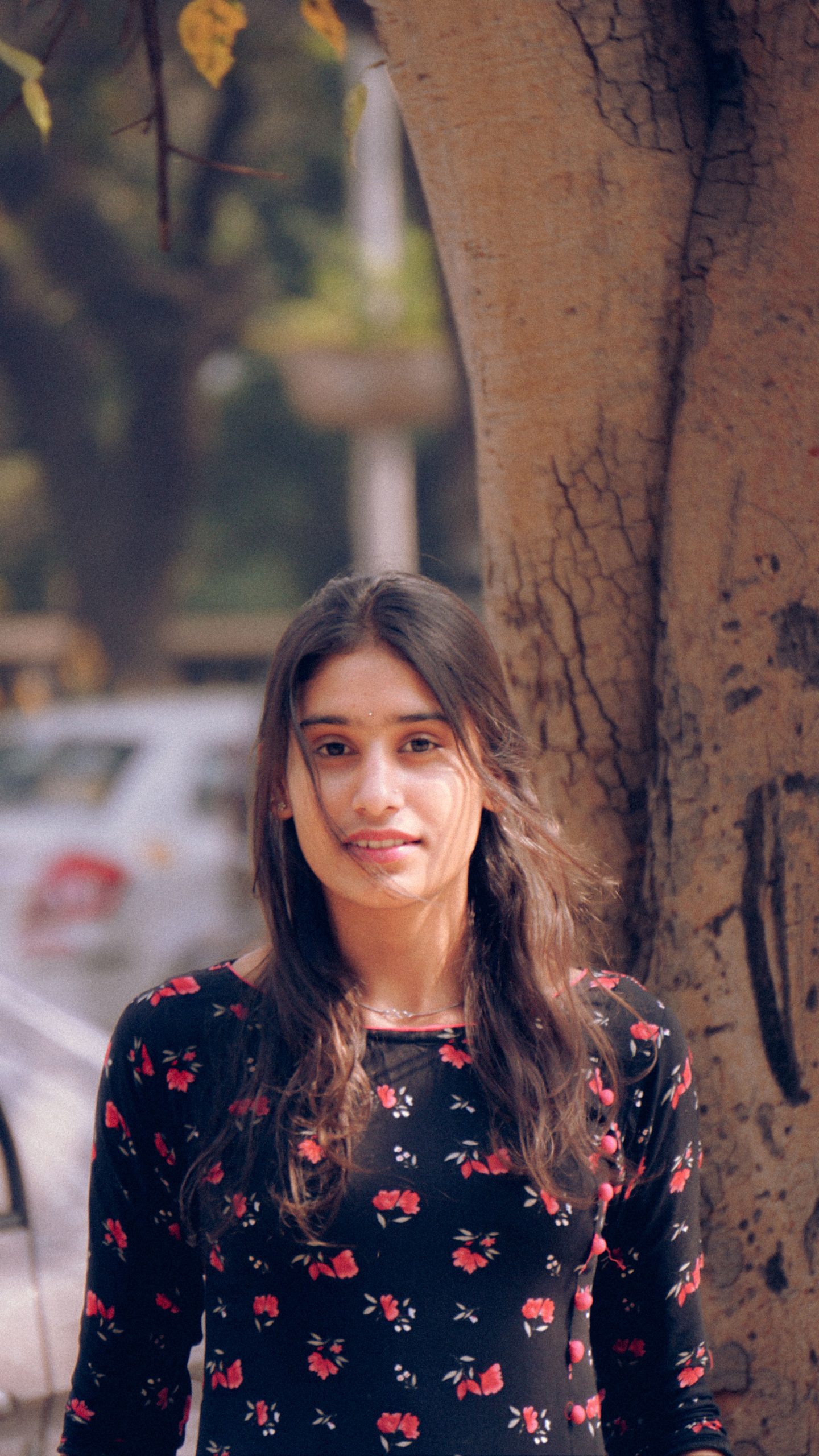 Portrait of a young woman with long brown hair, wearing a floral dress and posing by a tree.