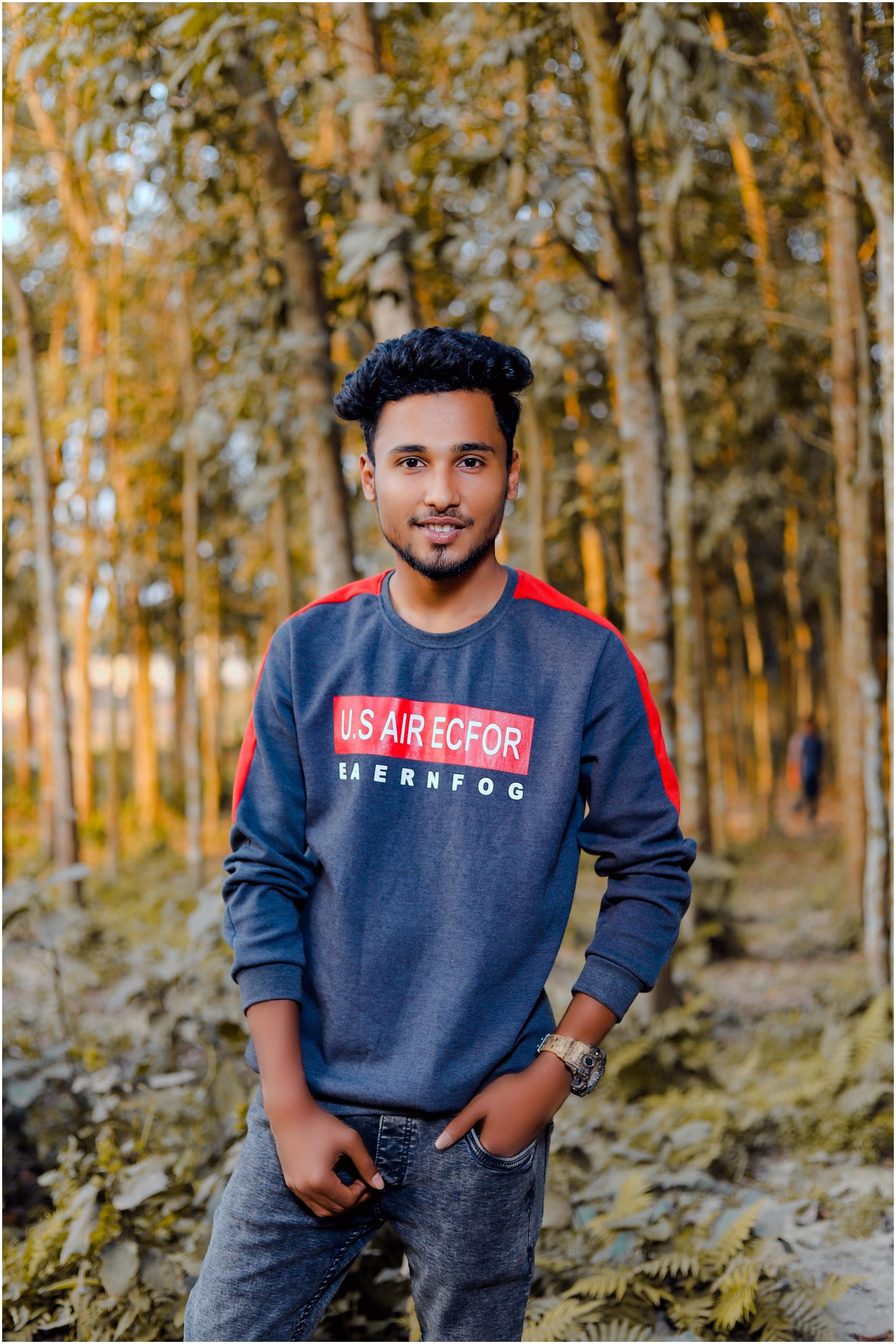 A young man smiling casually in a vibrant forest setting, wearing a sweatshirt.